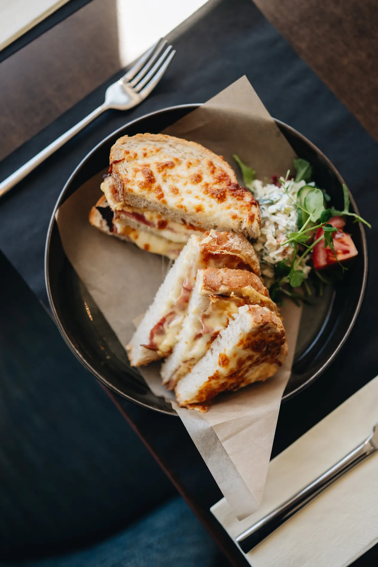Bovenaanzicht van een goudbruin gebakken tosti met rijkelijk gesmolten kaas en ham, geserveerd met een frisse bijpassende salade op een donker bord.